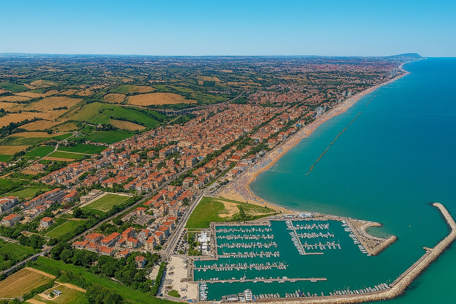 Vista aerea di Porto San Giorgio con il porto turistico, la spiaggia affollata e le colline marchigiane sullo sfondo, meta ideale per vacanze estive sulla costa adriatica.