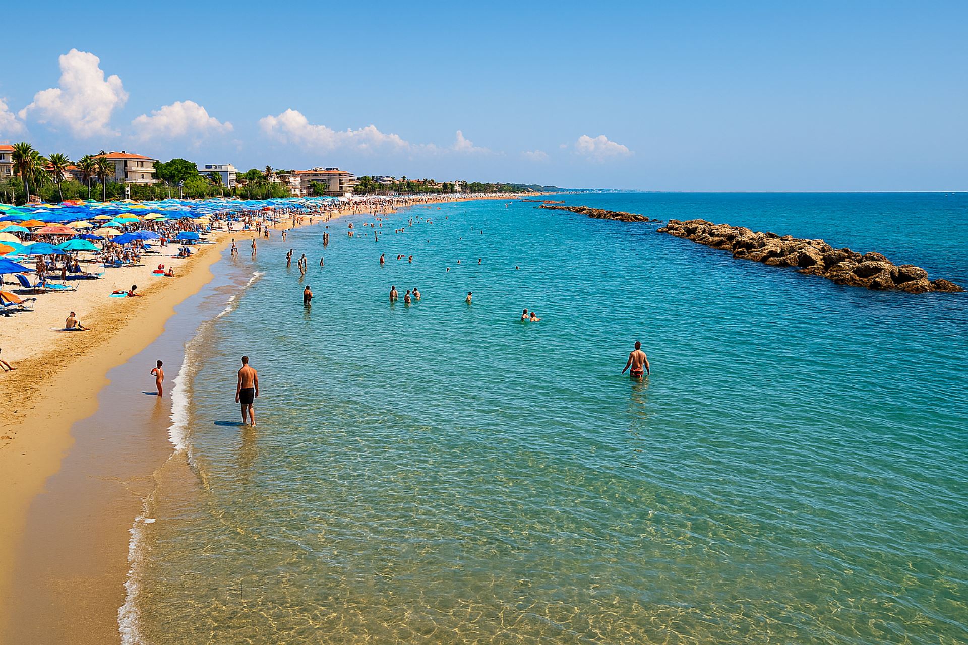 Panoramica estiva del litorale di Martinsicuro con mare calmo e trasparente, bagnanti in acqua e spiaggia affollata con ombrelloni colorati sullo sfondo urbano.